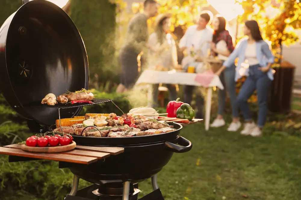 Outdoor backyard BBQ grill with food cooking and people gathered nearby.